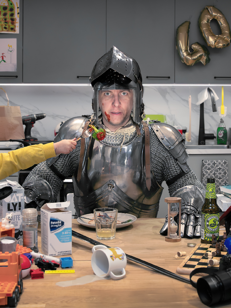 En man iklädd rustning från medeltiden sitter vid matbordet i ett modernt kök. Vardagskaos pågår runtomkring honom.