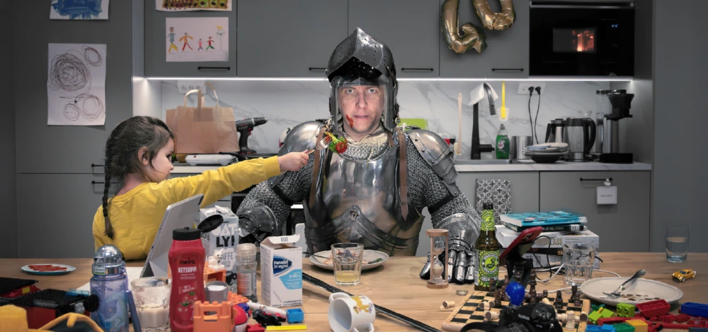 En man iklädd rustning från medeltiden sitter vid matbordet i ett modernt kök. Vardagskaos pågår runtomkring honom.