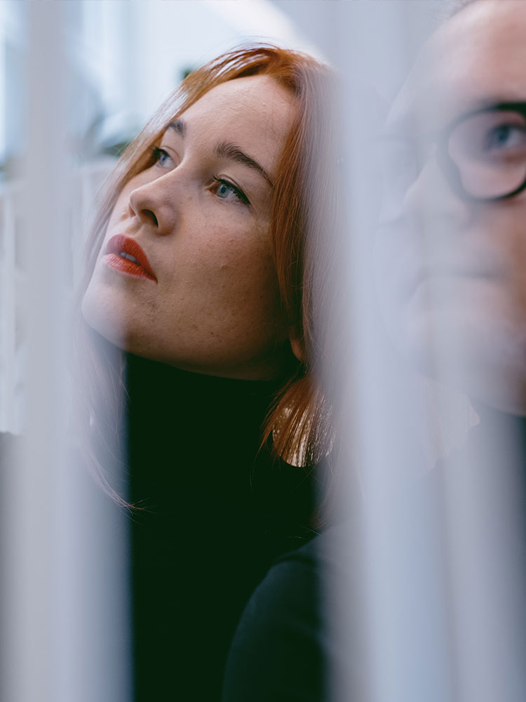 En kvinna och en man tittar snett uppåt. Framför dem fladdrar två skira, vita gardiner.