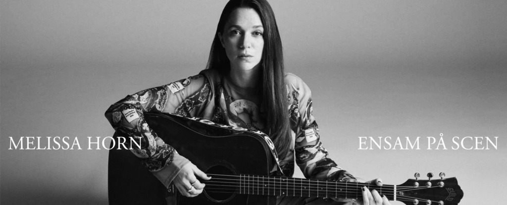 Svartvitt foto där Melissa Horn sitter ensam på golvet med en gitarr i famnen.