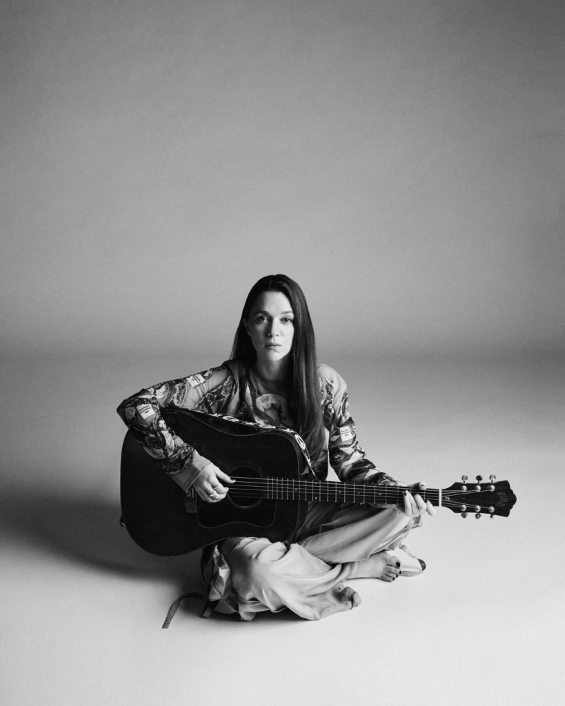 Svartvitt foto där Melissa Horn sitter ensam på golvet med en gitarr i famnen.