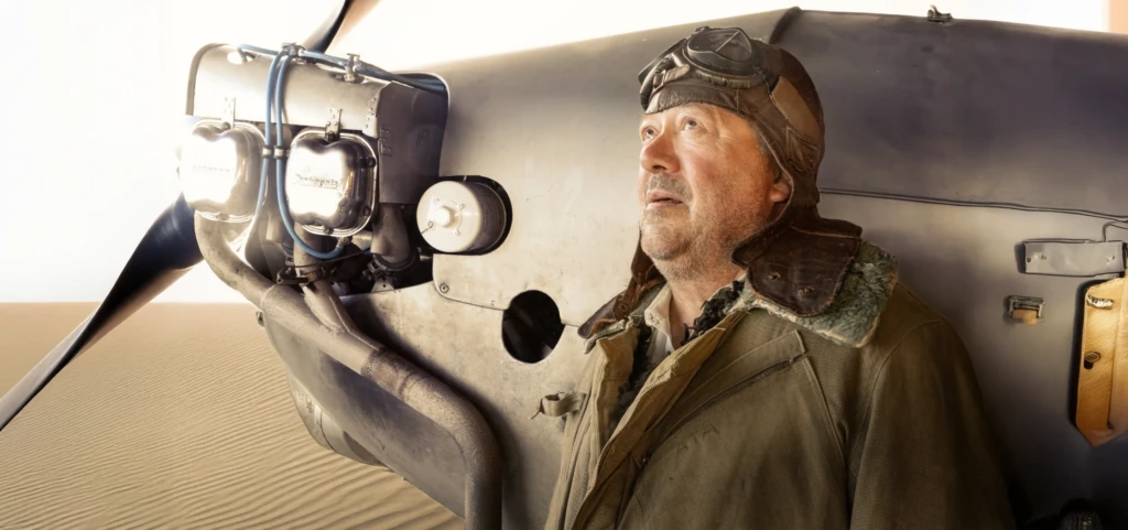 En man i gammaldags flygarjacka och pilotmössa står bredvid ett flygplan i en öken. Han ser upp mot himlen med ett eftertänksamt uttryck. Solens ljus reflekteras mot flygplanets metall. Reklambild för pjäsen Den lille prinsen. Mies vanhanaikaisessa lentäjän takissa ja lentäjänlakin kanssa seisoo lentokoneen vieressä autiomaassa. Hän katsoo ylöspäin mietteliäänä, ja auringon valo heijastuu koneen metallipinnasta. Mainoskuva näytelmästä Pikku prinssi. A man wearing a vintage pilot jacket and leather aviator cap stands beside an airplane in the desert. He looks upward thoughtfully as sunlight reflects off the plane’s metallic surface. Promotional image for the play The Little Prince.