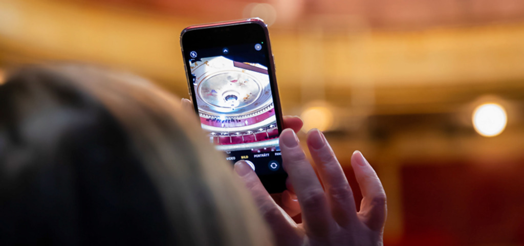 En person fotograferar teaterns salong med sin mobiltelefon. På skärmen syns takmålningen och kristallkronan i Åbo Svenska Teater. Finska: Henkilö ottaa valokuvan teatterisalista matkapuhelimellaan. Näytöllä näkyy Åbo Svenska Teaterin kattomaalaus ja kristallikruunu. English: A person takes a photo of the theatre auditorium with their phone. The screen shows the ceiling painting and chandelier of Åbo Svenska Teater.