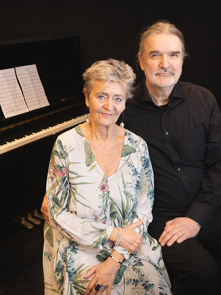 Två personer sitter bredvid varandra framför ett piano med noter uppställda på notstället. Personen i blommig klänning ler mot kameran medan personen i svart skjorta sitter tätt intill i en varm och stillsam porträttbild. Kaksi henkilöä istuu vierekkäin pianon edessä, jonka nuottitelineellä on avoimet nuotit. Kukkakuvioiseen mekkoon pukeutunut henkilö hymyilee kameralle, kun taas mustapaitainen henkilö istuu vieressä lämpimässä ja rauhallisessa muotokuvassa. Two people sit side by side in front of a piano with sheet music displayed on the stand. The person in a floral dress smiles toward the camera while the person in a black shirt sits close beside them in a warm and calm portrait.