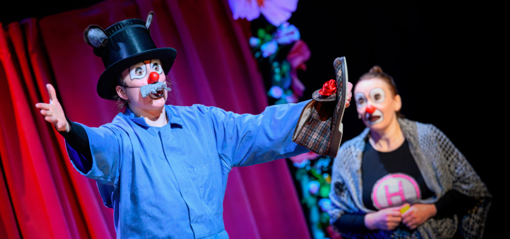 Två personer med clownsmink står på scen framför röda ridåer under ett livligt framträdande. Den ena personen gestikulerar med utsträckta armar och håller en hatt, medan den andra ser på i bakgrunden. Kaksi klovnimeikattua henkilöä esiintyy lavalla punaisten verhojen edessä vauhdikkaassa kohtauksessa. Toinen elehtii kädet levällään ja pitää hattua kädessään, kun taas toinen seuraa tapahtumaa taustalla. Two people wearing clown makeup perform on stage in front of red curtains during an energetic scene. One person gestures with outstretched arms while holding a hat, as the other watches in the background.